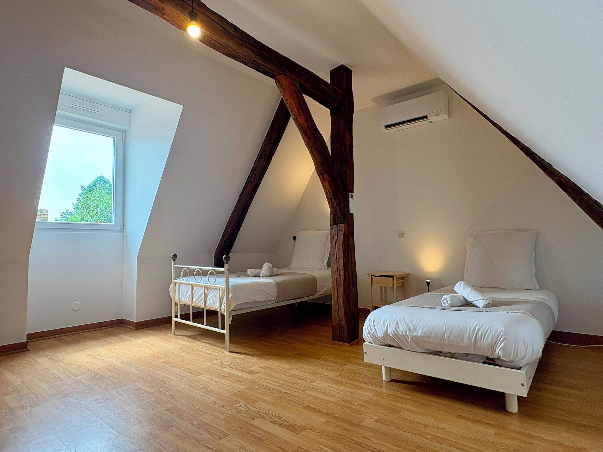 A room with a sloped ceiling, a window with a view of greenery, a bed, a small table, and a wall-mounted air conditioner.