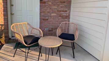 A patio with two wicker chairs and a small round table, a white door, and a brick wall.