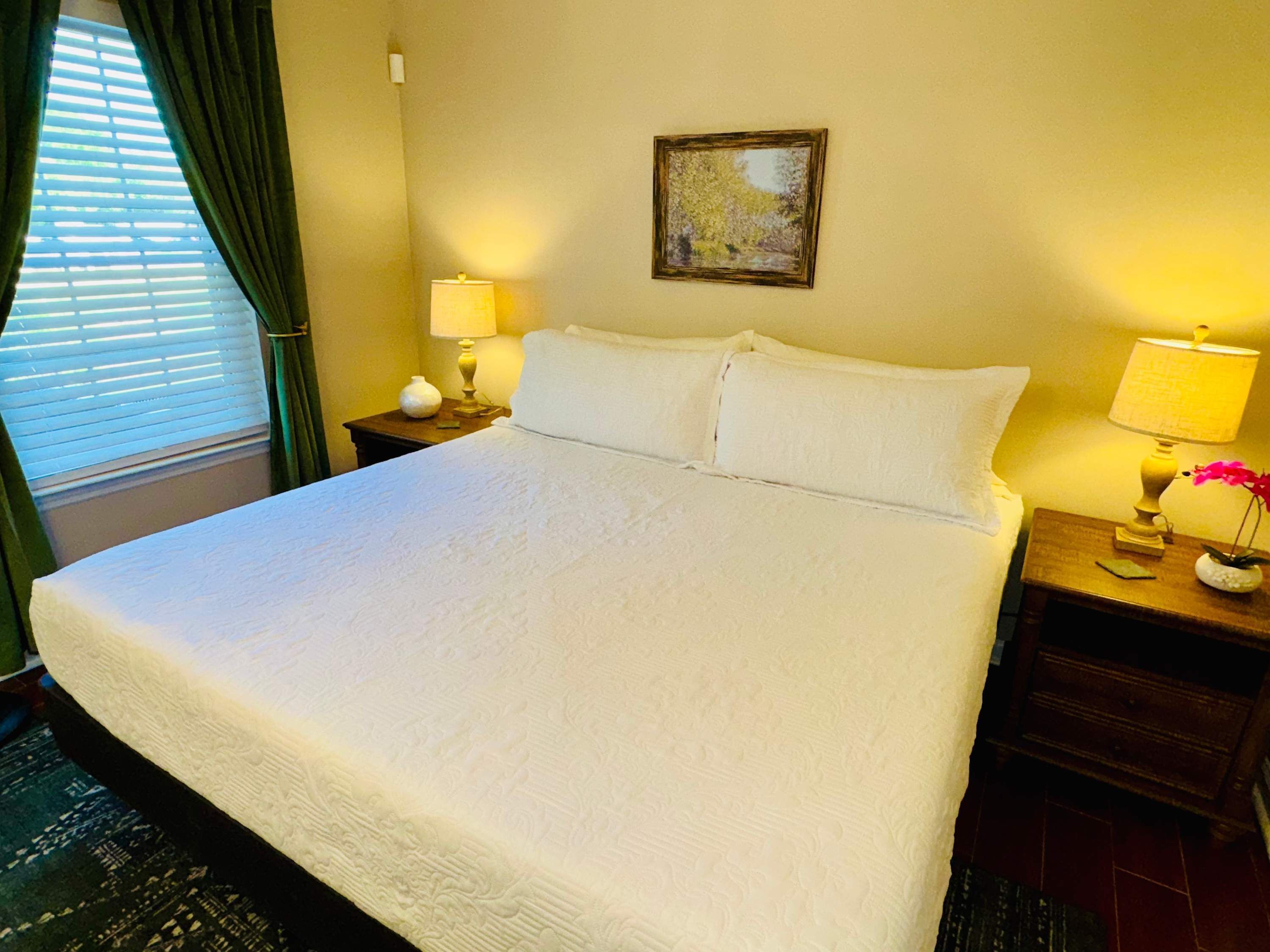 A neatly made bed with white linens, two bedside lamps, a small vase with flowers, and a framed picture on the wall.