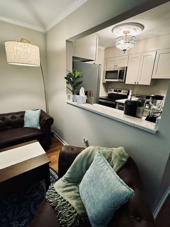 A modern living room with a brown leather sofa, a small table, and a mirror above a kitchen counter. - Lakefront Condo - King Bed Full Kitchen (Auburn)