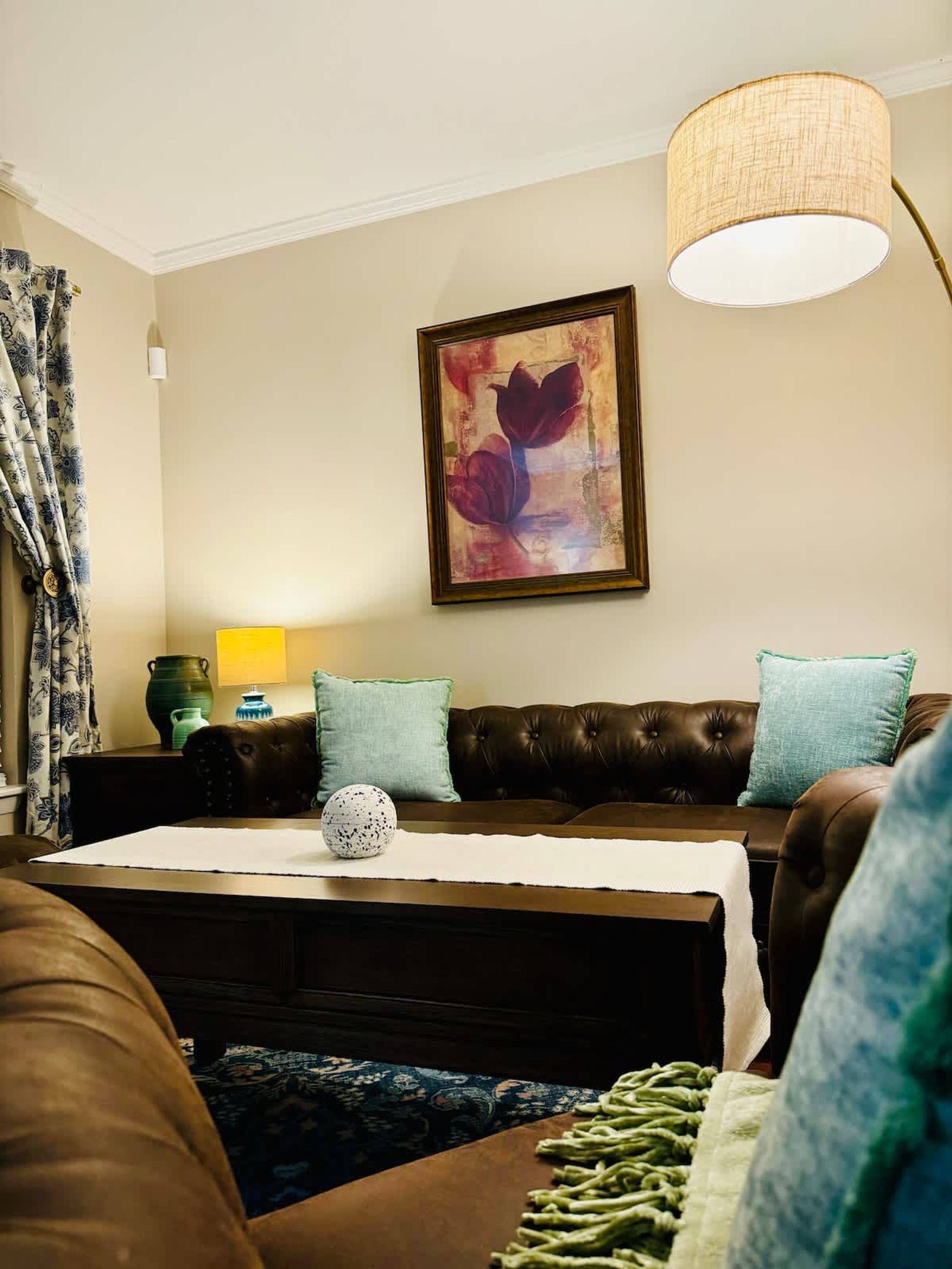 Una sala de estar con un sofá de cuero marrón, una mesa con mantel blanco, un jarrón decorativo y un cuadro en la pared.
