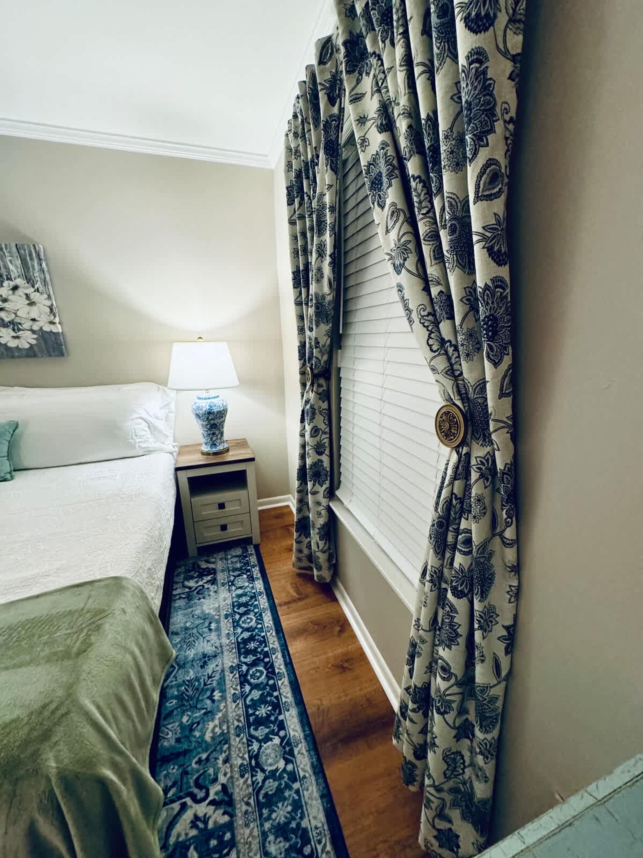 A bedroom with a bed, a nightstand with a lamp, a blue patterned rug, and floral curtains.