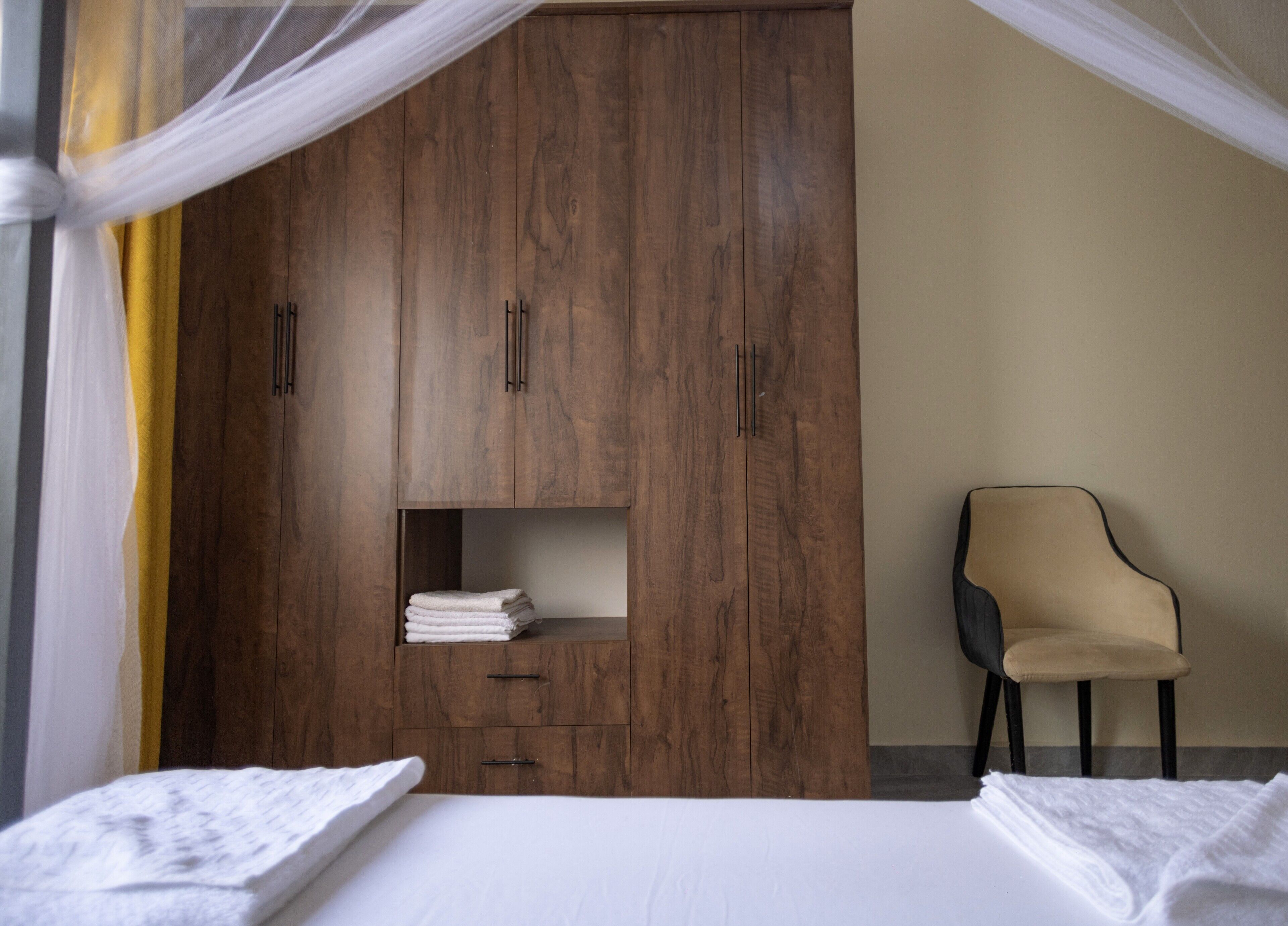 Une chambre avec une grande armoire en bois, une chaise et un lit recouvert de draps blancs.