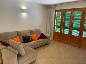 Ein Wohnzimmer mit einem beigen Sofa, einem kleinen Sessel und einer Holztür mit grünen Fensterläden.