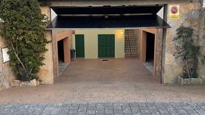 Ein überdachter Eingangsbereich mit einem Ziegeldach, eine Steinsäule mit einem Schild und ein gepflasterter Weg, der zu einem Gebäude mit grünen Fensterläden führt.
