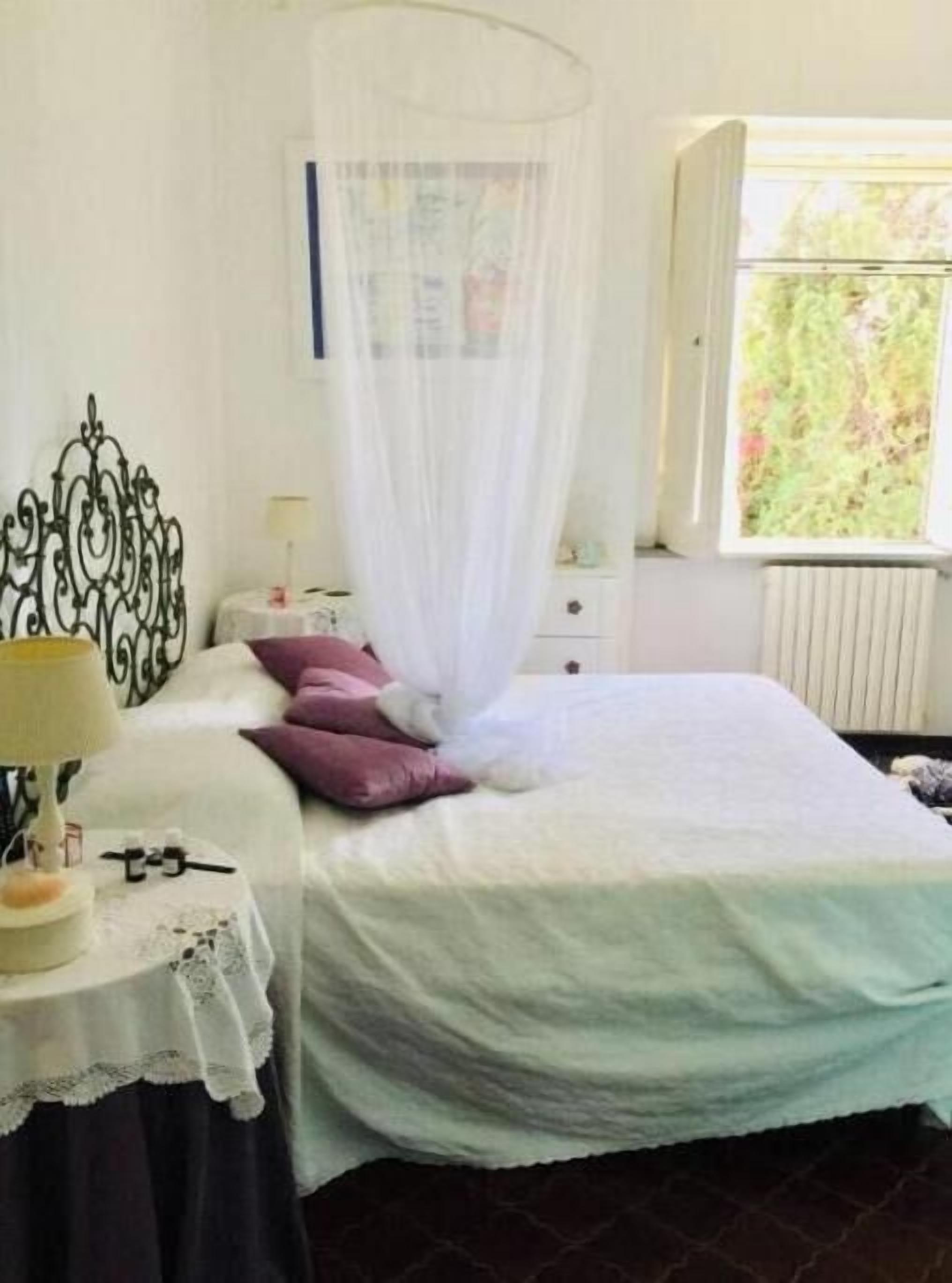 Ein Schlafzimmer mit Bett, Nachttisch mit Lampe, Fenster mit Blick auf Grünflächen und einem Vorhang.