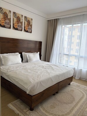 A bedroom with a wooden headboard, white bedding, a patterned rug, and artwork on the wall. - Serene 2-bedroom apartment perfect for your Nairobi getaway (Nairobi)