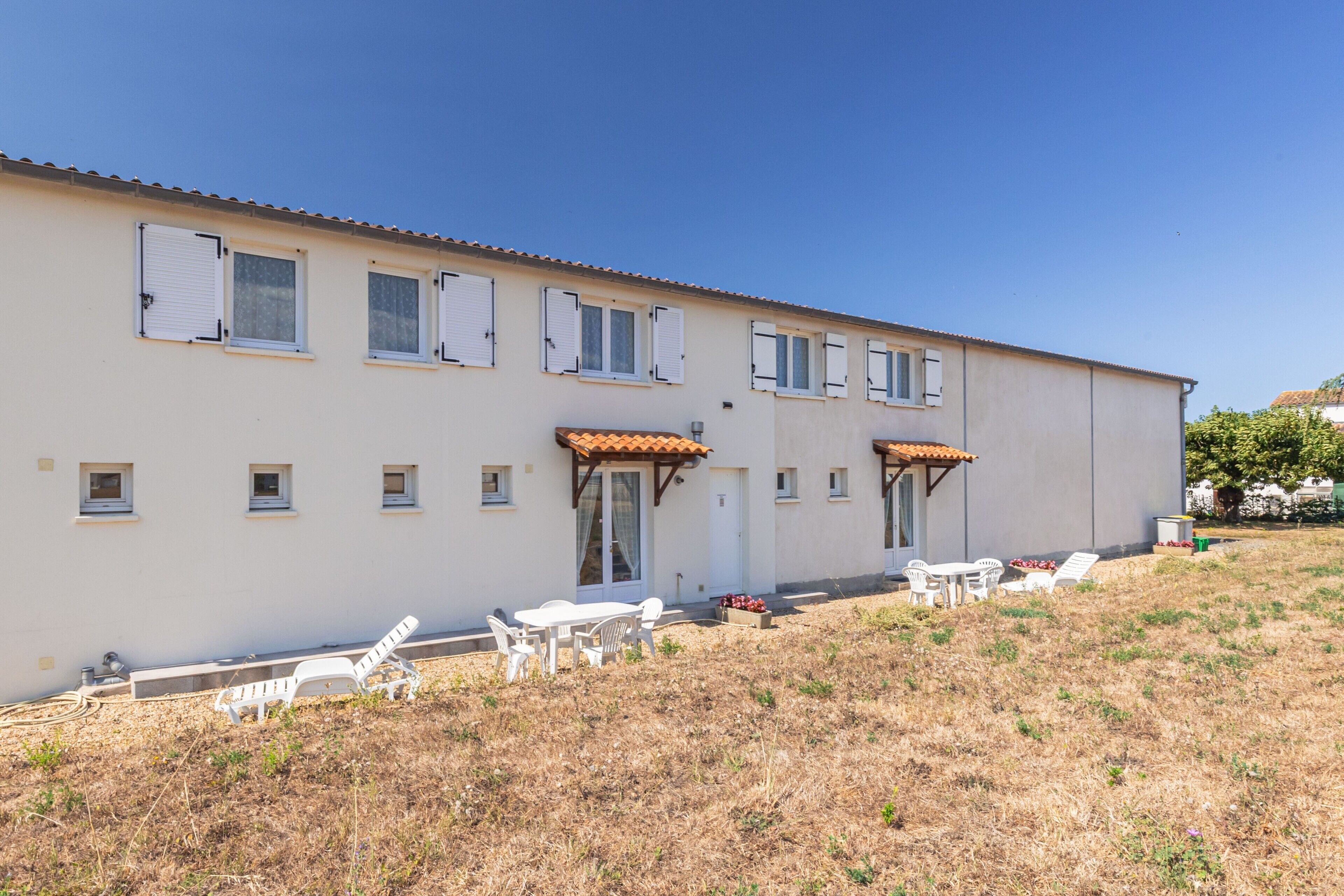 Un bâtiment à deux étages, doté d’un toit plat, de nombreuses fenêtres avec volets et d’un patio aménagé avec du mobilier d’extérieur.