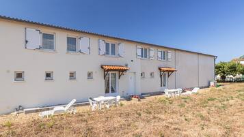 Un bâtiment à deux étages, doté d’un toit plat, de nombreuses fenêtres avec volets et d’un patio aménagé avec du mobilier d’extérieur.