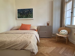 A bedroom with a bed, a nightstand, a chair, a painting, and a window. - Apartment 'Orange' in Bielefeld (Bielefeld)