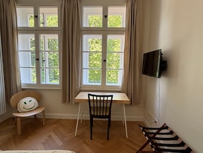 A room with a wooden floor, a desk with a chair, a television mounted on the wall, and a window with curtains. - Apartment 'Orange' in Bielefeld (Bielefeld)