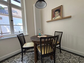 A dining area with a round table, two chairs, a pendant light, a radiator, a framed picture on the wall, and patterned floor tiles. - Apartment 'Orange' in Bielefeld (Bielefeld)
