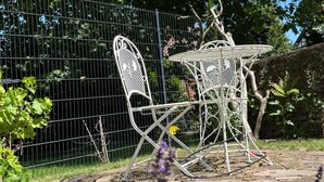 Un ensemble de table et de chaises en métal pour l’extérieur, avec un chemin en pierre, entouré de verdure et délimité par une clôture en métal.