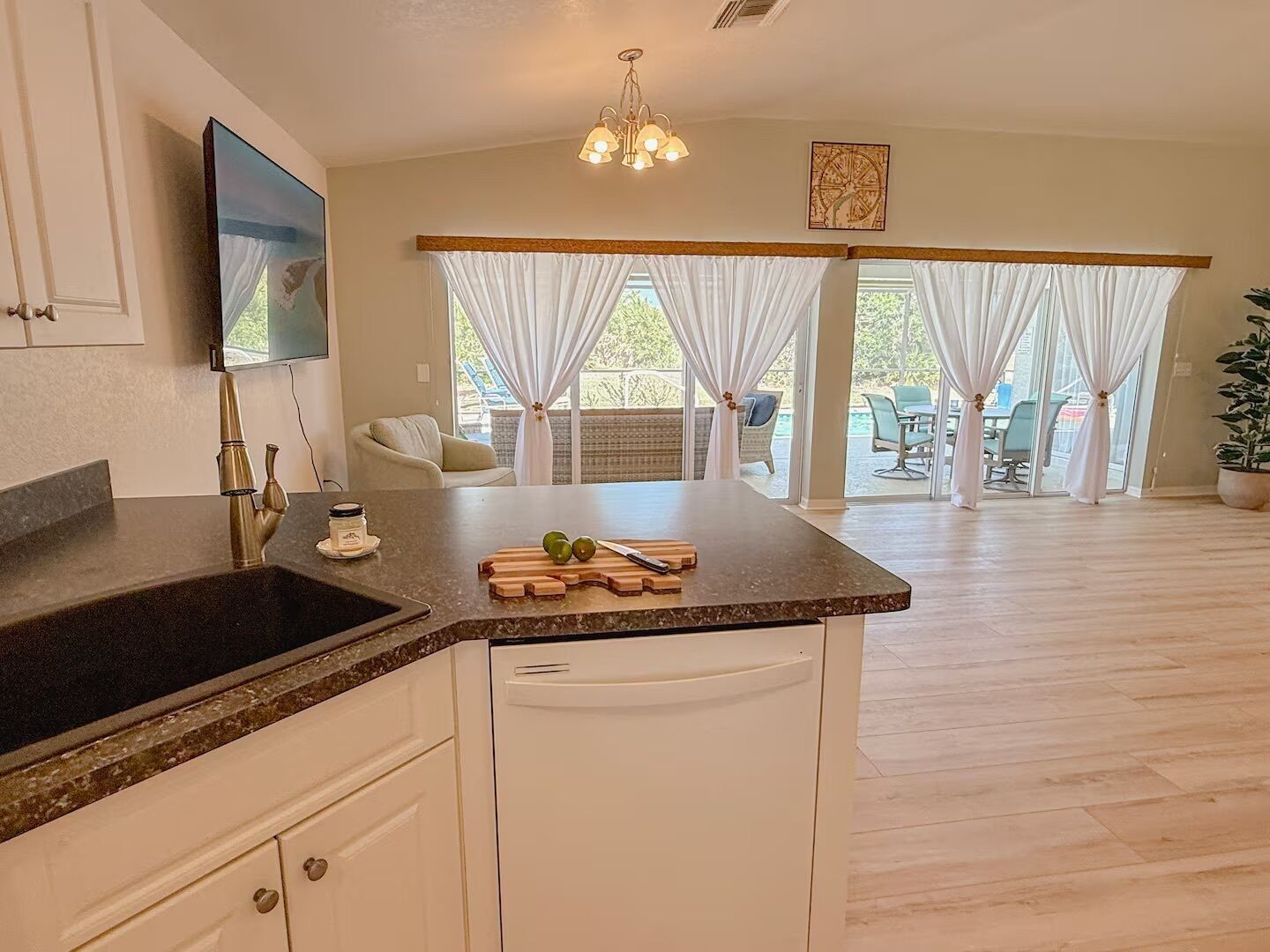 Cocina con encimera de granito, armarios blancos y lavavajillas integrado. Se ve una zona de comedor con mesa y sillas a través de ventanas grandes con cortinas blancas.