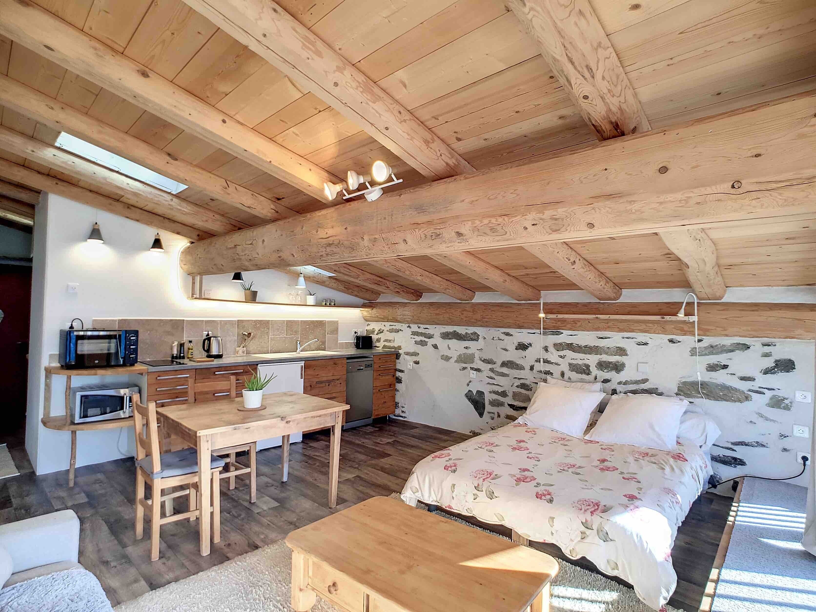 Ein rustikaler Raum mit Holzbalkendecke, einem Bett mit floralem Bettbezug, einem Holztisch, einer Küchenzeile mit Mikrowelle und einer Steinwand.