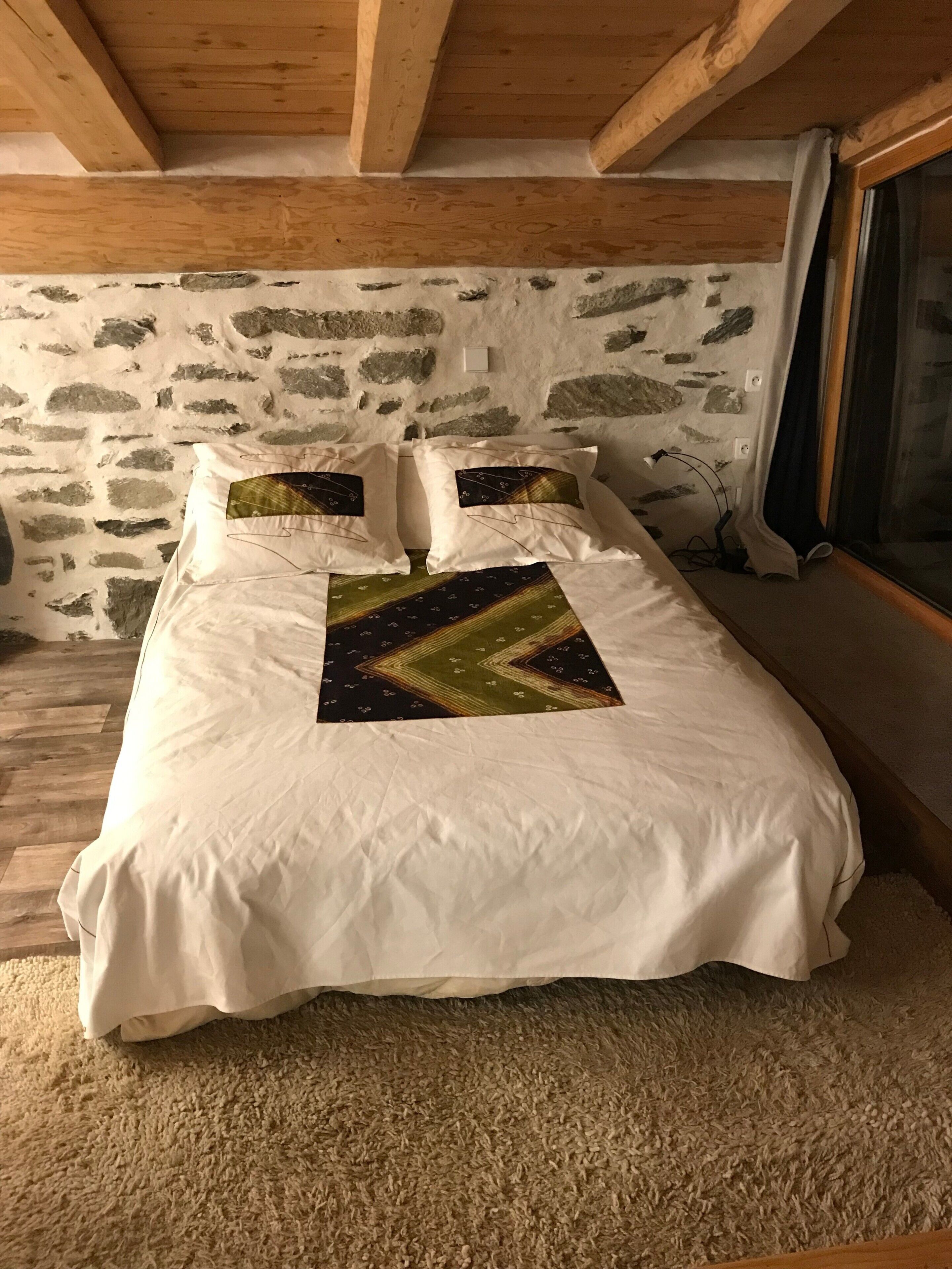 A bed with white bedding and a geometric-patterned pillow, set against a stone wall and wooden beams.