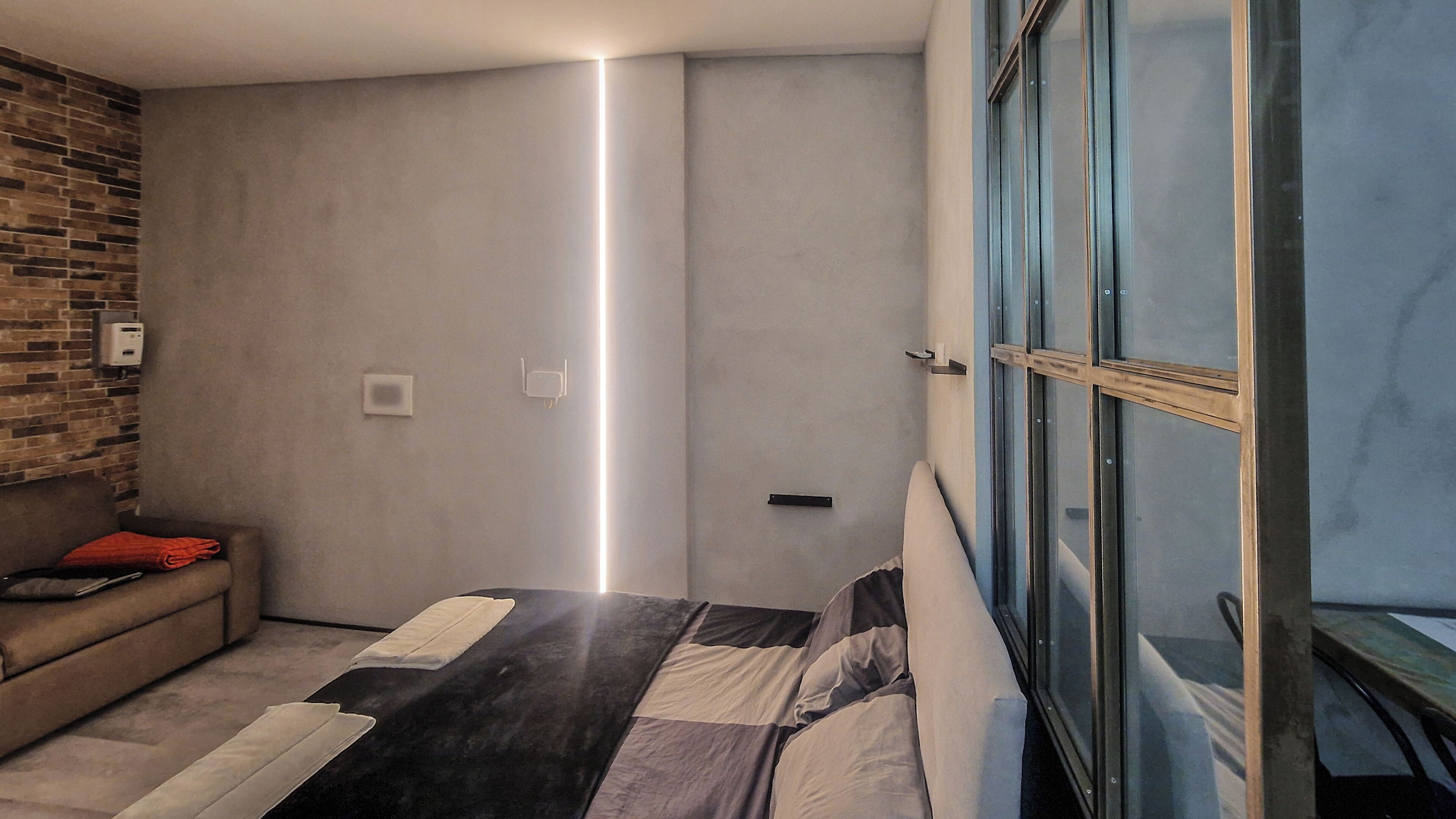 A modern bedroom with a bed, a sofa, a brick wall, and a large window.