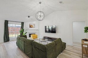 A modern living room with a sectional sofa, a yellow armchair, a fireplace, a flat-screen TV, and a large window with curtains. - Fireside Retreat| Sleeps 10 | Foosball | Fire pit (Grand Prairie)