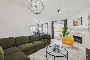 Een woonkamer met een hoekbank, een gele fauteuil, een ronde salontafel en een open haard.