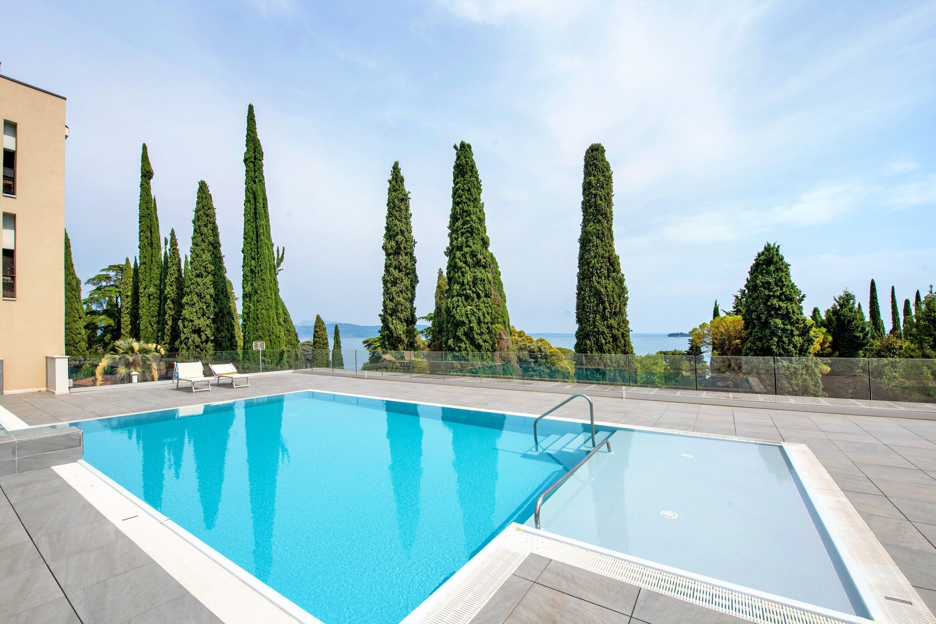 A rectangular swimming pool with a metal ladder, surrounded by a tiled deck, tall cypress trees, and a view of the sea.