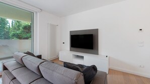 En moderne stue med et fladskærms-TV monteret på en hvid væg, en grå sofa og et vindue med udsigt til grønne omgivelser.