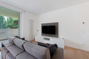 Ein modernes Wohnzimmer mit flachbildfernseher an einer weißen Wand, einem grauen Sofa und einem Fenster mit Blick ins Grüne.
