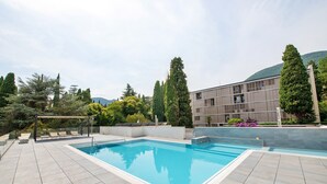 Et rektangulært svømmepool med fliseterrasse, omgivet af træer og en bygning med moderne arkitektur.