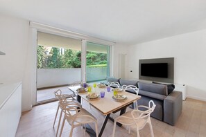 A modern living room with a dining area, a grey sofa, a flat-screen TV, and a large sliding glass door to a balcony. - Apartment 'Michela Flat in Gardone' with Shared Pool, Wi-Fi and Air Conditioning (Gardone Riviera)