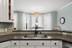 A kitchen with a double sink, white cabinetry, a hanging light fixture, and a view of a dining area with a blue tablecloth. - Beautiful-Lakefront Farmhouse Retreat w/ Pool (Summerton)