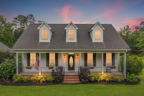 A two-story house with a front porch, a flag, and a well-maintained lawn. - Beautiful-Lakefront Farmhouse Retreat w/ Pool (Summerton)