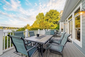 An outdoor patio with a table and chairs, a white railing, and a view of trees and a body of water. - Big Water Lodge | Hot Tub, Fire Pit, & Lake Views (Macon)
