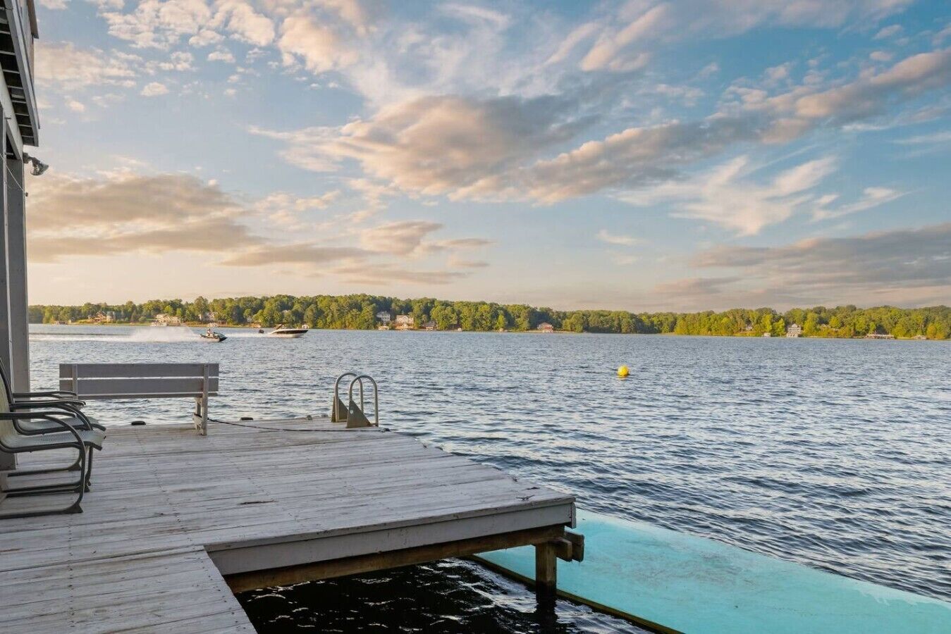 Une terrasse en bois avec un banc et une piscine qui s’étend jusqu’à un lac, le tout entouré d’arbres et sous un ciel nuageux.