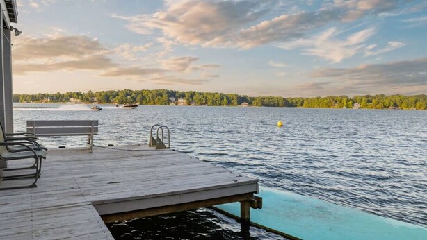 A wooden deck with a bench and a pool extending into a lake, surrounded by trees and a cloudy sky. - Big Water Lodge | Hot Tub, Fire Pit, & Lake Views (Macon)