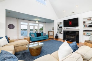 A spacious living room with a large TV, a fireplace, and a view through the windows. - Big Water Lodge | Hot Tub, Fire Pit, & Lake Views (Macon)