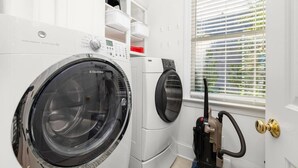 A laundry room with two front-loading washing machines, a dryer, and a vacuum cleaner. - Big Water Lodge | Hot Tub, Fire Pit, & Lake Views (Macon)