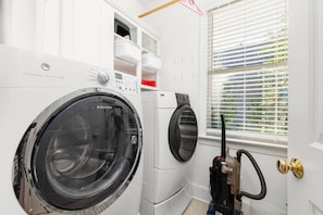 A laundry room with two front-loading washing machines, a dryer, and a vacuum cleaner. - Big Water Lodge | Hot Tub, Fire Pit, & Lake Views (Macon)