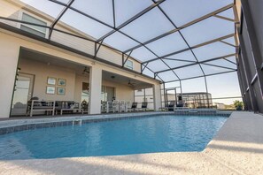 Une piscine entourée de parois en verre, avec un espace extérieur aménagé pour s’asseoir et une terrasse couverte.