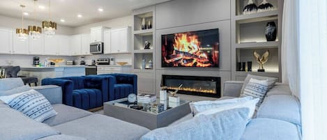 A modern living room with a fireplace, a television, and a kitchen area in the background.
