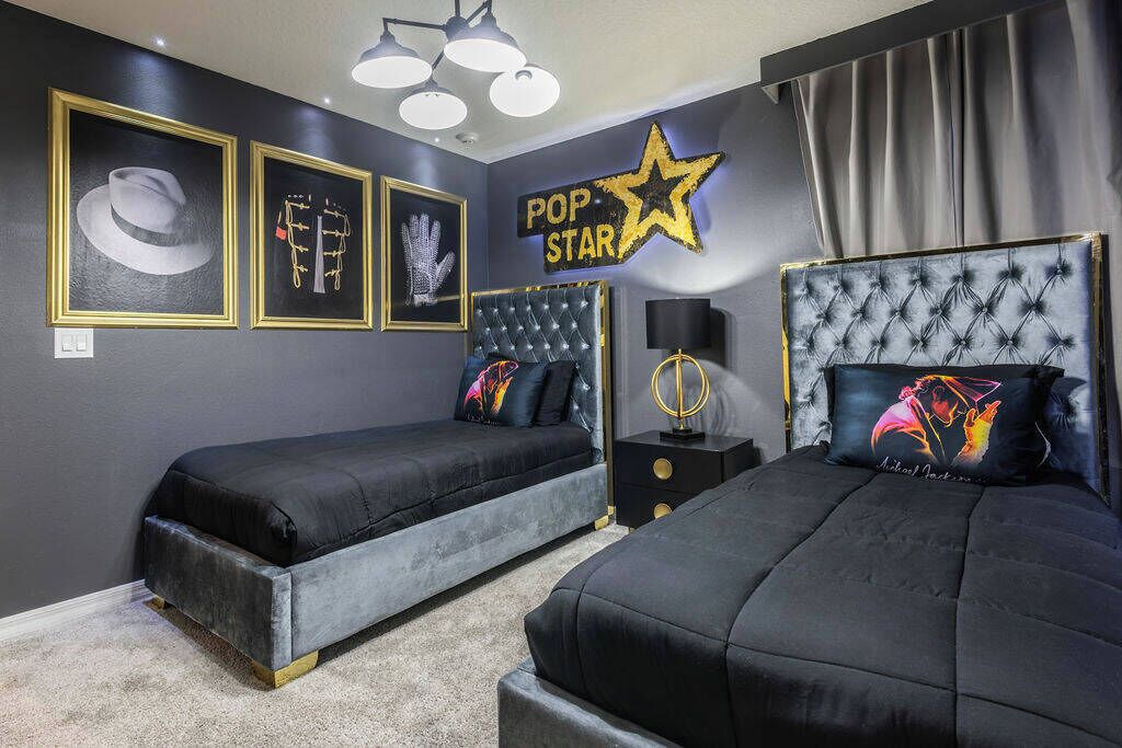 A room with two beds, a wall-mounted star sign, framed artwork, and a bedside table with a lamp.