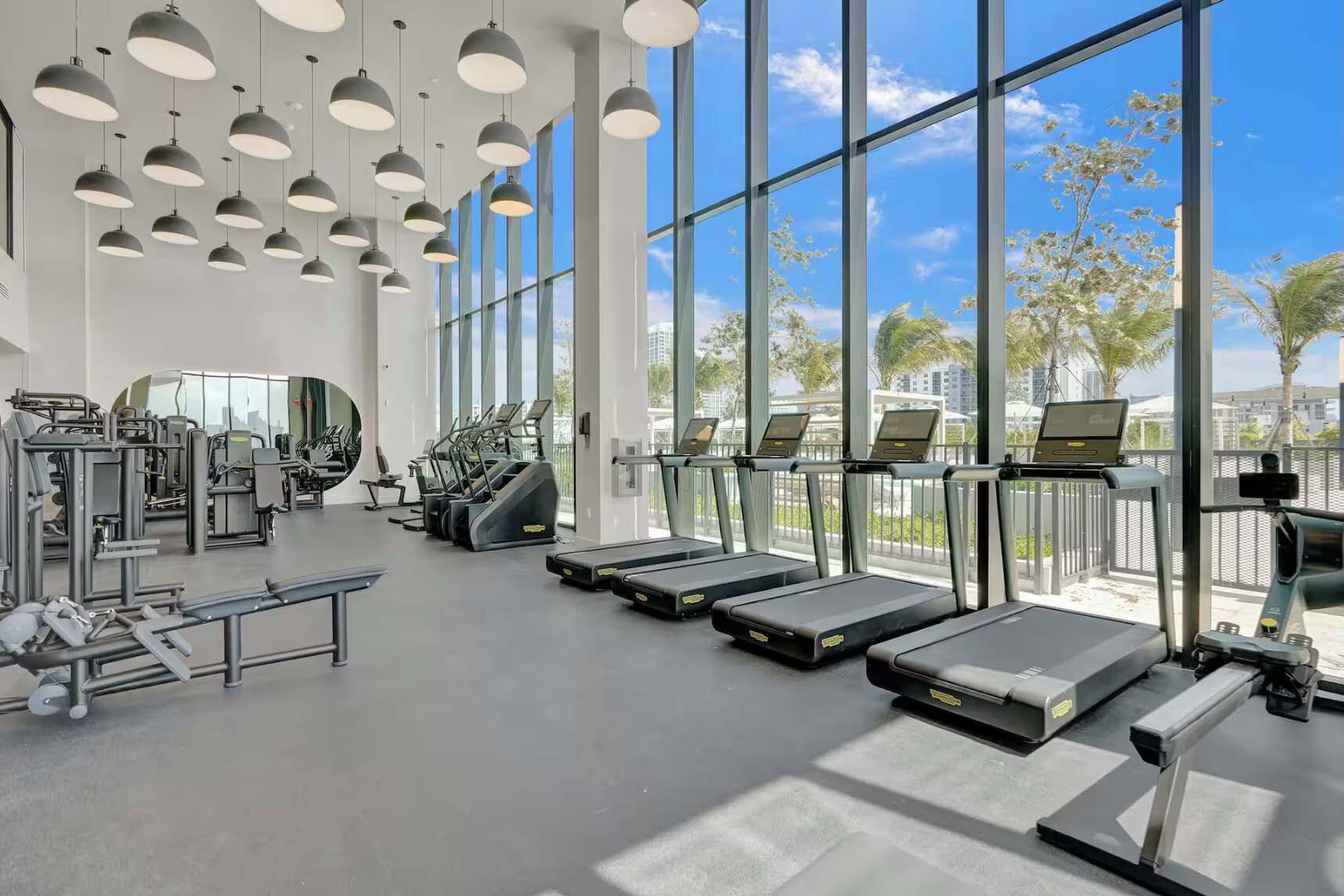 A gym with treadmills, weight machines, and large windows offering a view of palm trees and buildings.
