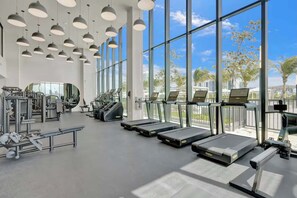 A gym with treadmills, weight machines, and large windows offering a view of palm trees and buildings. - Miami Beach Luxury Stay Balcony Sea Views & Pool (Miami Beach)