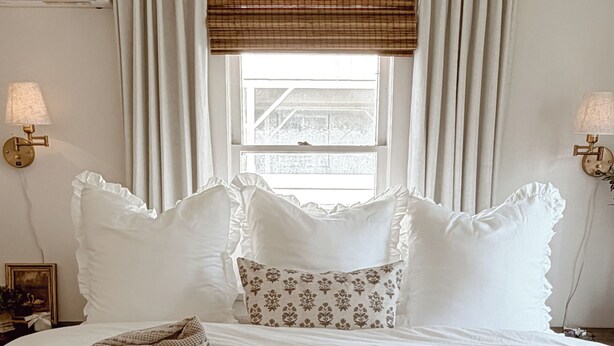 Un lit bien fait, avec du linge de lit blanc et des coussins décoratifs, une couverture à motifs et une fenêtre avec des stores.