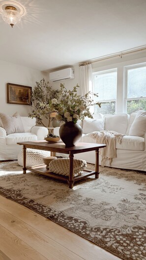 A living room with a white sofa, a wooden coffee table with a vase of flowers, a patterned rug, and a painting on the wall. - Charming 1940s cottage near college and downtown (Caldwell)
