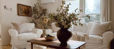 Una sala de estar con dos sofás blancos, una mesa de centro de madera con un florero y una alfombra estampada, además de un cuadro en la pared.