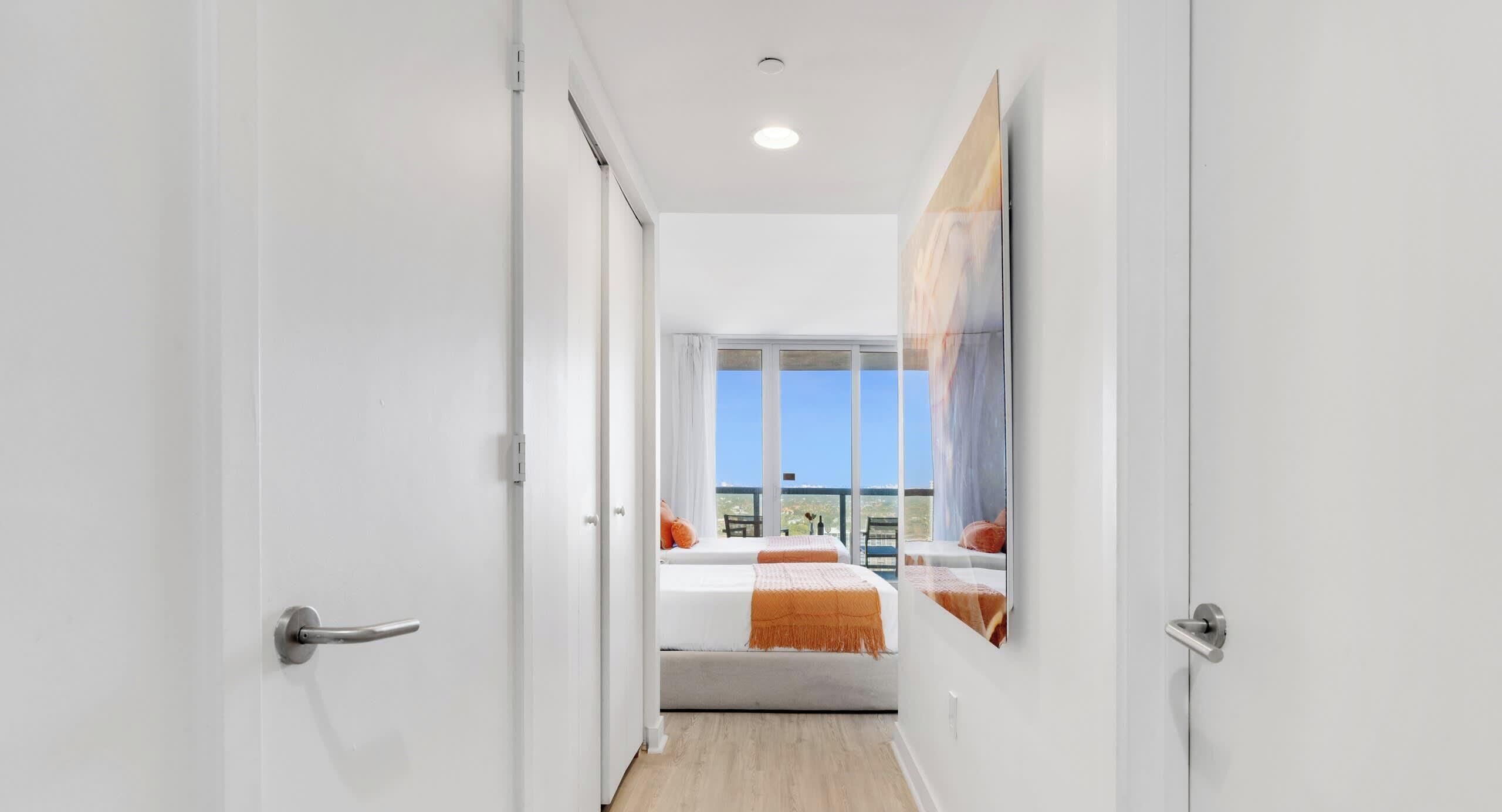 Ein enger Flur führt zu einem Schlafzimmer mit einem großen Fenster, einem Bett mit orangefarbener Bettwäsche und einem Bild an der Wand.