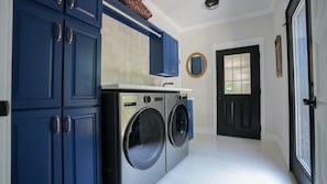Ein Waschraum mit zwei Waschmaschinen mit Frontlader, einem blauen Schrank mit Messinggriffen, einem weiß gefliesten Boden und einer schwarzen Tür.