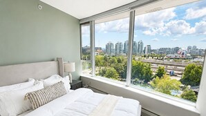 Een slaapkamer met een groot bed, een nachtkastje, een lamp en een stadszicht door een groot raam.
