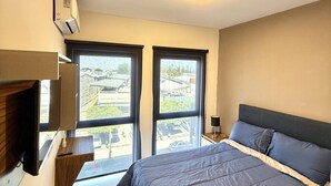 Un dormitorio con cama, dos ventanas, una lámpara de techo y un televisor sobre un mueble.