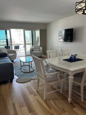 A modern living room with a sofa, dining table, chairs, and a TV mounted on the wall. - Newly remodeled Beachfront 2BR-2Bath Condo / Pool & Gulf Views (Fort Myers Beach)