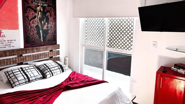 A bedroom with a bed, a TV, a window with a view, and a red cabinet. - Penthouse with ocean view, one block from the beach, with private garage. (Florianópolis)